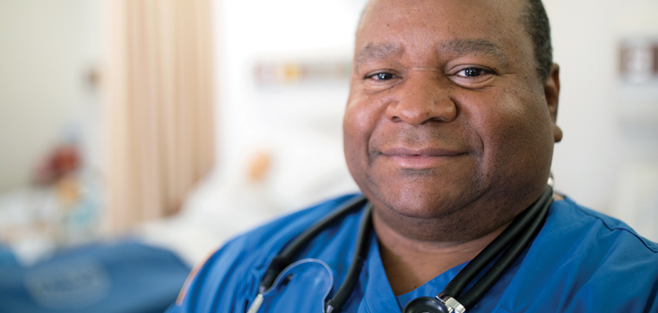 man in blue scrubs and a stethoscope around his neck with a patient in bed in the background