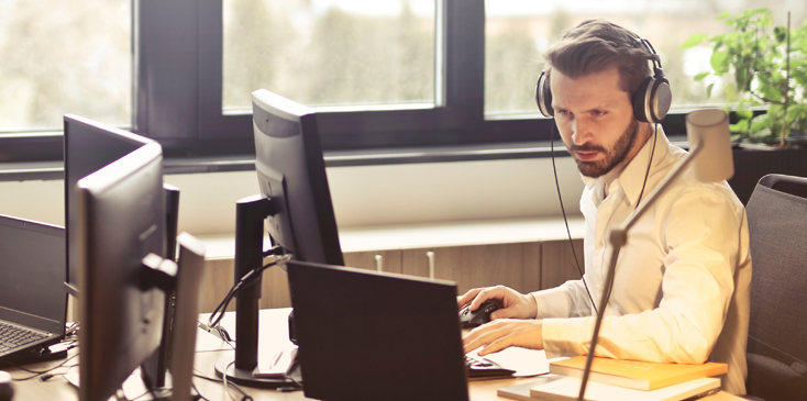 man with headphones sits at a desk working on a laptop and monitor