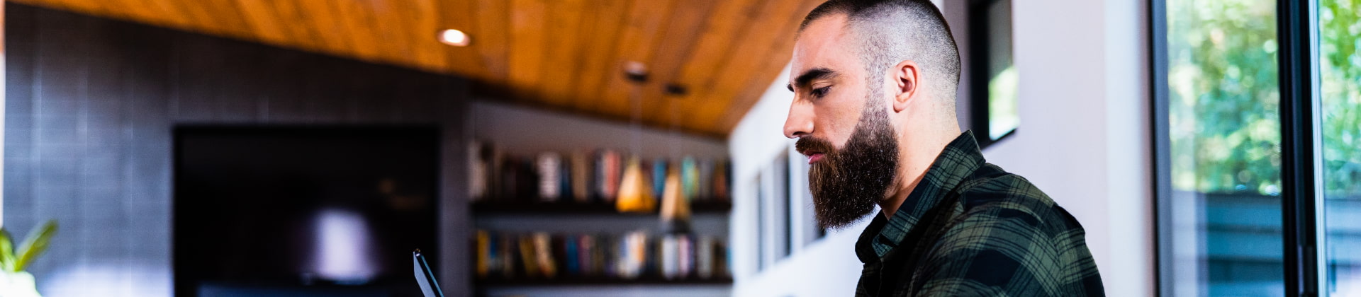 Man with a beard and wearing a dark flannel shirt works inside on a laptop