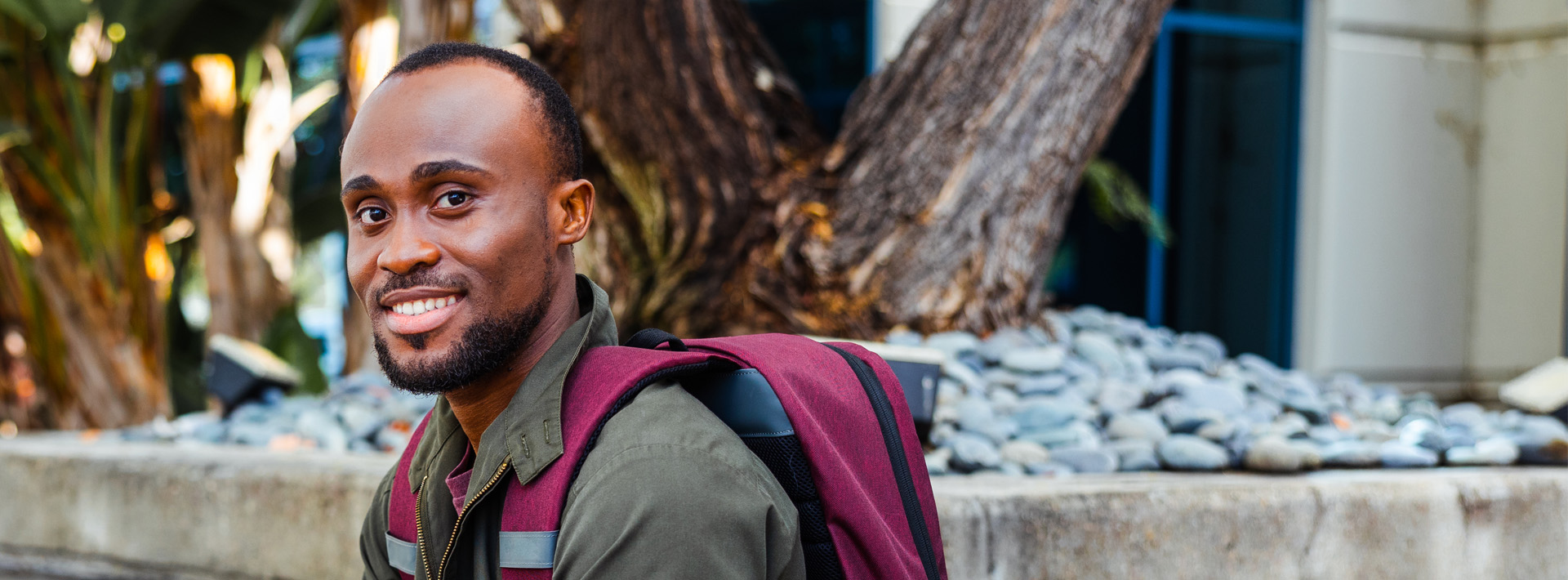 A man wearing a burgundy backpack sits in front of a tree outside