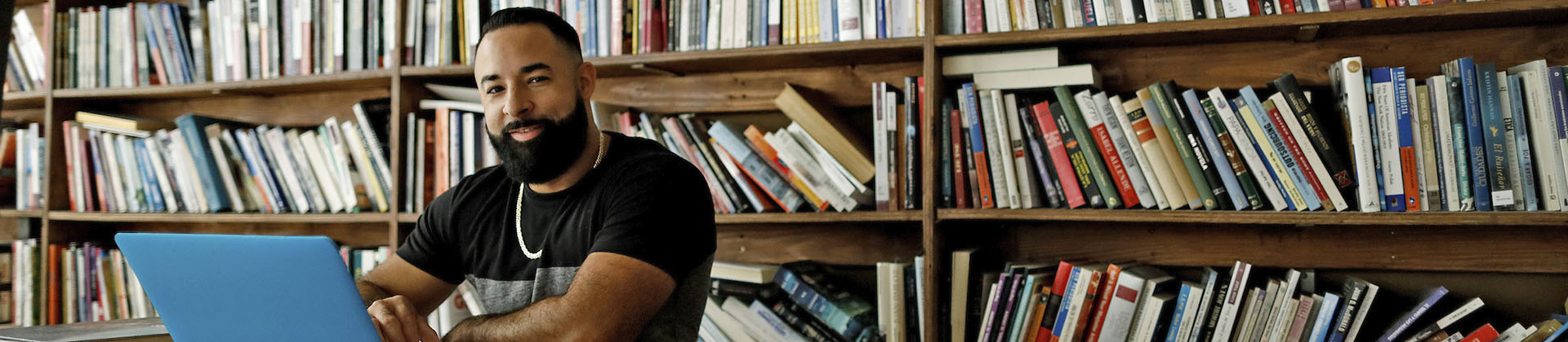 Man with a beard in a t-shirt sits in library on a laptop