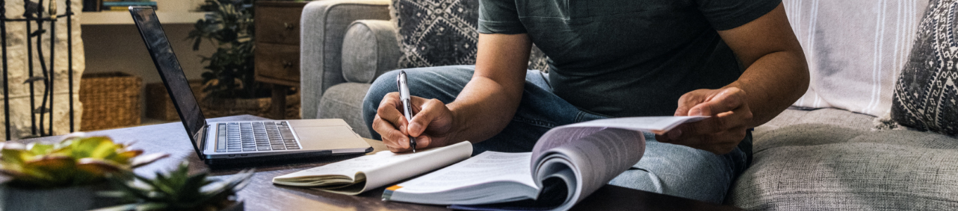 man sitting on couch with laptop, textbook, and notebook on coffee table as he takes notes