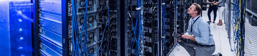 man kneeled down in server room looking up at rack holding tablet