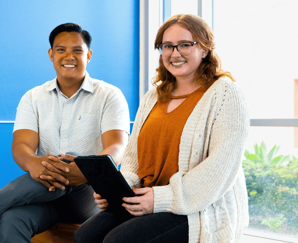 two students sit at a window. one of them is holding a tablet computer